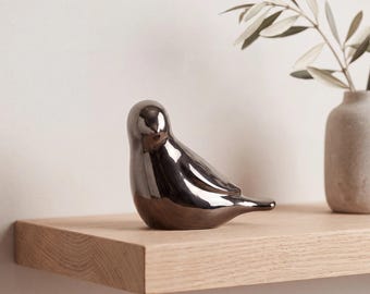 May include: A smooth, reflective metallic silver bird figurine with a rounded body and a pointed tail. The object features a polished, mirror-like finish that captures light, showcasing a simple, stylized avian form in a seated position.