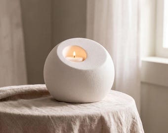 May include: A spherical white candle holder with a matte, textured finish. The object features a circular opening on the upper side, containing a small, lit white tea light candle with a visible flame, casting a warm glow from within the center.