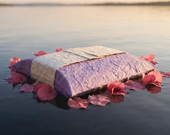 May include: A rectangular, textured lavender paper package wrapped with a wide, cream-colored paper band, floating on water and surrounded by scattered pink bougainvillea flower petals, creating a soft, layered visual composition.