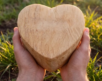 May include: A heart-shaped object carved from light-colored wood with a visible natural grain pattern. The surface shows subtle texture and markings from the carving process, presenting a smooth, rounded form held in two hands.