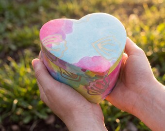 May include: A heart-shaped box featuring a watercolor-style pattern in shades of pink, blue, and yellow, accented with gold-colored line drawings of seashells. The lid and base are aligned, showcasing a smooth, rounded exterior surface.