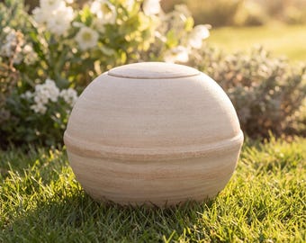 May include: A spherical, light beige stone urn with a smooth, matte finish and a horizontal seam encircling the center. The object features a rounded lid on top and a solid, rounded base, presenting a clean, uniform, and neutral appearance.