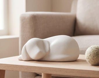 May include: A smooth, matte white ceramic sculpture of a dog in a resting, curled-up position. The figurine features rounded, simplified forms representing the head, ears, and body of the animal, presented as a solid, singular piece.