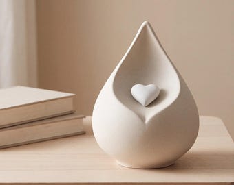 May include: A teardrop-shaped ceramic vessel in a matte off-white finish, featuring a smooth, concave front surface with a small, raised white heart centered within the hollow. The object has a solid, rounded base and a pointed top.
