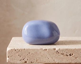 May include: A smooth, rounded, periwinkle blue ceramic object with a glossy finish. The piece has an irregular, pebble-like shape with soft, curved edges and a solid, uniform color throughout its surface, reflecting light on its top.