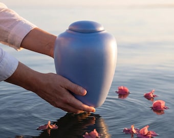 May include: A smooth, matte periwinkle blue cremation urn with a rounded lid and a tapered body. The vessel features a solid, minimalist design with a soft finish, held securely in place above the water's surface.