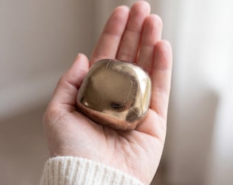 May include: A small, smooth, rounded square object with a metallic bronze finish. The surface has a reflective, polished appearance with subtle texture variations across its curved edges and flat top, creating a solid, compact form.
