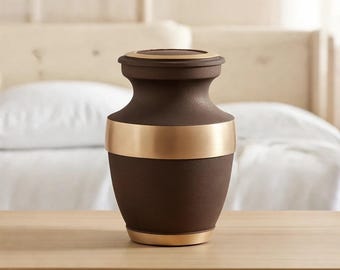 May include: A dark brown urn with a textured matte finish, featuring a wide, polished gold-tone band around the center and a matching gold-tone rim on the lid and base. The vessel has a classic tapered neck and a rounded body shape.