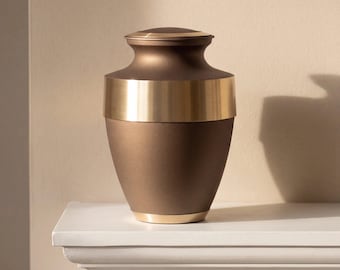 May include: A metal cremation urn featuring a matte bronze finish on the body and lid, accented by a wide, polished gold-tone band around the upper section and a thin gold-tone ring at the base. The vessel has a tapered neck and lid.