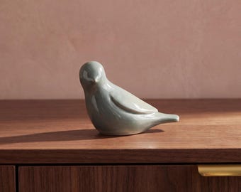 May include: A smooth, light gray ceramic bird figurine with a glossy finish. The bird is depicted in a resting position with its head turned slightly to the side, featuring a rounded body, a small beak, and a short, pointed tail.