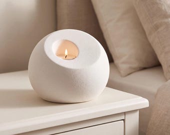 May include: A spherical white candle holder with a textured matte finish and a circular opening at the top. A small lit tea light candle is placed inside the recessed center, casting a warm glow from the flame within the white stone-like object.