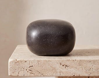 May include: A smooth, rounded, dark charcoal gray stone object with a speckled texture. The object has a solid, pebble-like form with a polished finish, featuring subtle light gray flecks distributed across its dark, matte-like surface.