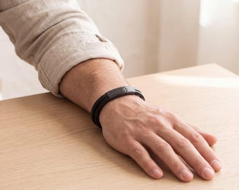 May include: A dark brown braided leather bracelet featuring a smooth, matte-finish rectangular magnetic clasp. The bracelet consists of multiple strands woven together to create a textured appearance, secured by a solid, minimalist metal closure.