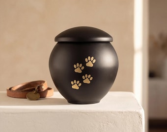 May include: A matte black spherical pet urn featuring a lid and four gold-colored paw print designs on the front. Beside the urn sits a brown leather collar with a circular metal tag attached to the buckle hardware.