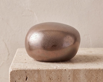May include: A smooth, rounded, pebble-shaped object with a metallic bronze finish. The surface has a subtle, speckled texture and a reflective sheen, creating a solid, compact form that rests evenly on a flat, textured surface.