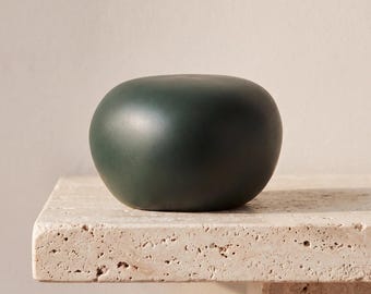 May include: A smooth, rounded, dark forest green object with a matte finish. The item has a solid, pebble-like shape with no visible markings, textures, or patterns on its surface. It is a singular, compact, and uniform decorative piece.