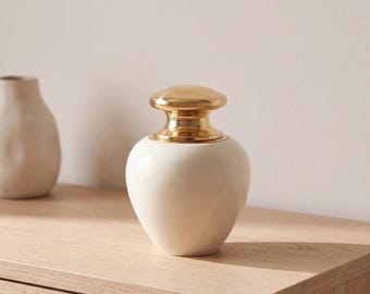 May include: A cream-colored ceramic jar with a smooth, rounded body and a polished, reflective gold-toned metal lid. The lid features a wide, flat top and a tapered neck that sits securely on the opening of the vessel.