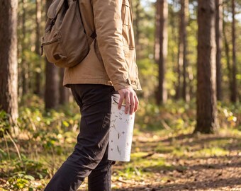 May include: A cylindrical white water bottle featuring a delicate botanical pattern of small green leaves and brown stems, held by a person wearing a tan hooded jacket, a brown backpack, and dark trousers while walking through a wooded forest area.