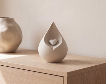 May include: A teardrop-shaped beige ceramic urn featuring a small, centered heart-shaped emblem. Beside it sits a larger, rounded, irregular beige ceramic vessel. Both objects have a matte, textured finish and neutral tones.
