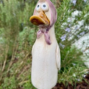 Könnte beinhalten: Ein skurriler Gartenstecker mit einer Keramikente mit überraschtem Gesichtsausdruck. Die Ente hat ein hellrosa Kopftuch und einen braunen Schnabel. Der Körper ist weiß mit gesprenkelten Details, perfekt für den Garten.