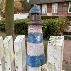 Könnte beinhalten: Eine blaue und weiße Keramik-Leuchtturmfigur mit braunen Details, die auf einem weißen Lattenzaun sitzt.