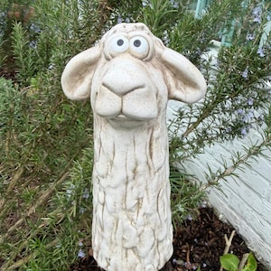 Könnte beinhalten: Ein cremefarbener Schaf-Gartenstecker mit einer strukturierten, geschnitzten Oberfläche. Das Schaf hat große, ausdrucksstarke Augen und ein detailliertes Gesicht. Der Stecker steht in einem Garten mit grünem Laub im Hintergrund und verleiht dem Garten Charme.
