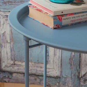 Könnte beinhalten: Ein runder Beistelltisch in hellblau mit einem Metallgestell und einem Buch mit floralem Muster darauf. Der Tisch steht vor einer blau-weiß gestrichenen Wand.