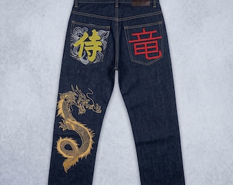 May include: Dark blue denim jeans featuring a gold embroidered dragon on the left leg. The back pockets display Japanese kanji characters; the left pocket has a gold character with white line art, and the right pocket has a red character.