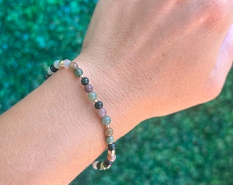 May include: A bracelet made of small, round beads in shades of green, brown, black, and white. The beads are strung on a gold chain.