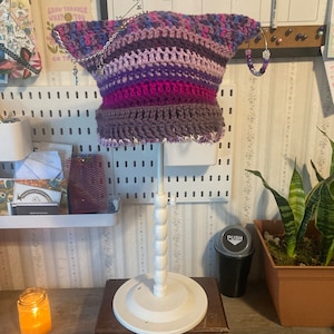 May include: A purple, pink, and gray crocheted hat with a silver chain and beaded embellishments. The hat is displayed on a white stand.