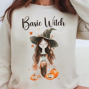 Könnte beinhalten: Ein beiger Pullover mit dem Aufdruck "Basic Witch" auf der Vorderseite. Der Pullover zeigt eine Comic-Illustration einer Hexe mit einem schwarzen Hut und einem Kürbis.