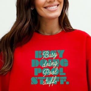 May include: A red sweatshirt with the text "Busy doing Pool stuff" in teal and yellow lettering. The sweatshirt is a solid red color and has a crew neck.
