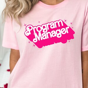 Puede incluir: Camiseta rosa con el texto "Program Manager" en una fuente retro rosa y blanca.