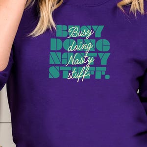 May include: A purple sweatshirt with the text "Busy doing Nasty stuff" in teal and white. The text is arranged in a stacked format. The sweatshirt has long sleeves and a crew neck.