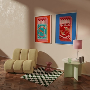 May include: Two framed prints with a blue and orange background featuring a box of matches with a tiger design. The text on the prints reads "You are my perfect match" and "The perfect match fire". The prints are hanging on a light beige wall above a light brown couch and a green table with a pink lamp.