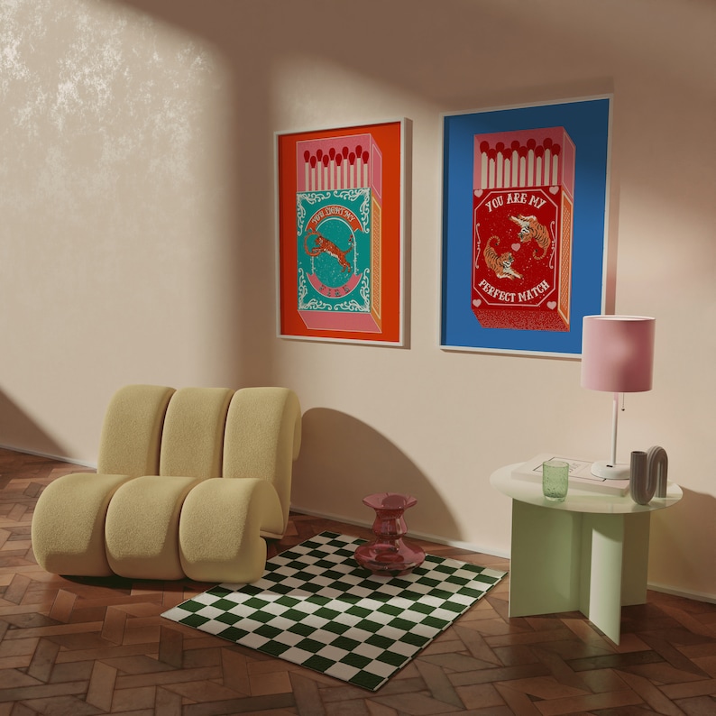 May include: Two framed prints with a blue and orange background featuring a box of matches with a tiger design. The text on the prints reads "You are my perfect match" and "The perfect match fire". The prints are hanging on a light beige wall above a light brown couch and a green table with a pink lamp.