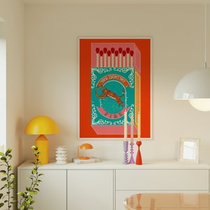 May include: A colorful illustration of a matchbox with a tiger and the text "You light my fire". The matchbox is on a red background and there are three candlesticks on a white shelf below the image.