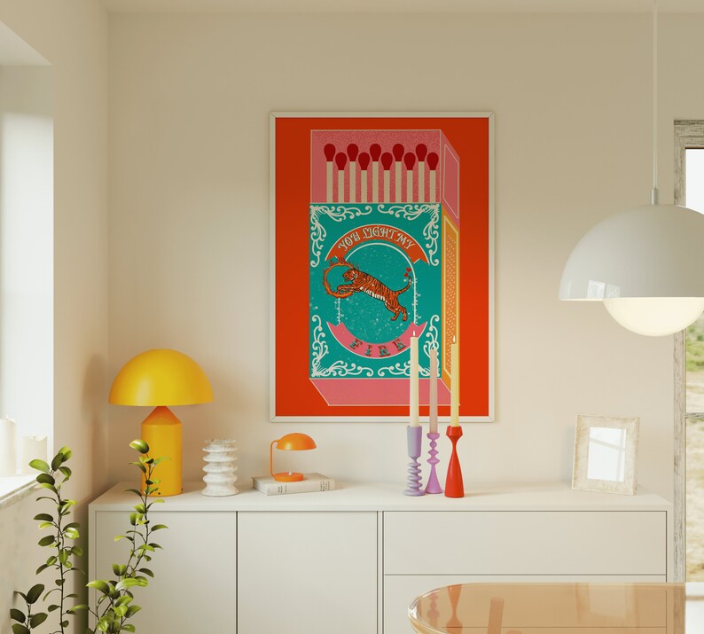 May include: A colorful illustration of a matchbox with a tiger and the text "You light my fire". The matchbox is on a red background and there are three candlesticks on a white shelf below the image.