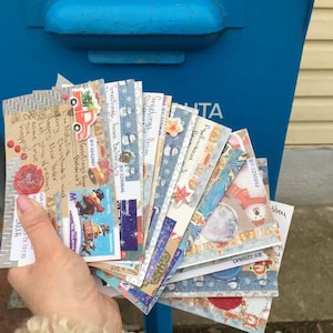 May include: A hand holds a collection of Christmas cards with festive designs, stamps, and handwritten messages. The cards feature images of snowmen, Christmas trees, and other holiday motifs. A blue postbox is in the background.