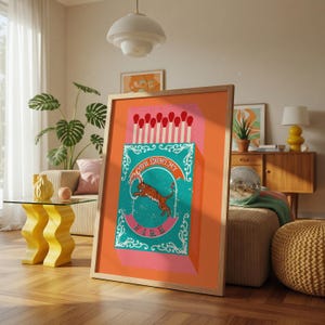May include: A framed art print featuring a matchbox design with the text "YOU LIGHT MY FIRE". The matchbox is teal and pink, with a tiger illustration. The matches are red and white. The artwork is displayed in a living room setting.