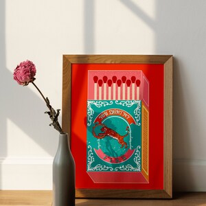 May include: A framed illustration of a matchbox with a tiger on the front. The matchbox is red with a teal and gold design. The text on the matchbox reads "You light my fire".