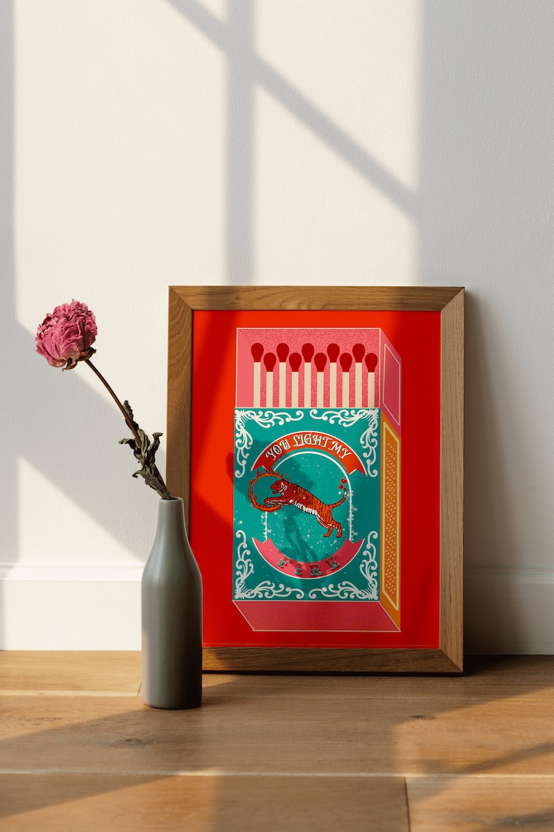 May include: A framed illustration of a matchbox with a tiger on the front. The matchbox is red with a teal and gold design. The text on the matchbox reads "You light my fire".