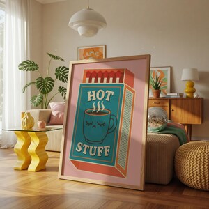 以下が含まれることがあります： 「HOT STUFF」の文字と、目を閉じたターコイズ色のマグカップが描かれたマッチ箱のデザインの額入りアートプリント。ピンク色の背景と木製のフレームで、インテリアに最適です。