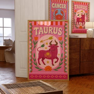 May include: Three framed art prints featuring zodiac signs. The largest print displays "TAURUS" with a pink bull illustration, surrounded by flowers and an arrow. Two smaller prints show "CANCER" with a crab and another "TAURUS" print. All are in wooden frames.