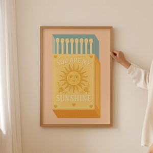 May include: A framed art print featuring a matchbox design with a smiling sun illustration. The text "YOU ARE MY SUNSHINE" is displayed on the matchbox. The color palette includes shades of yellow, orange, and blue.