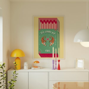 May include: A green and white illustration of a box of matches with the text "You Light My Fire" and two tigers facing each other. The illustration is framed and hanging on a wall above a white dresser with a yellow lamp and candlesticks.