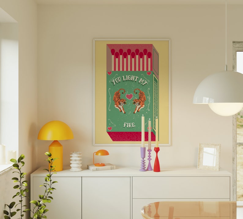 May include: A green and white illustration of a box of matches with the text "You Light My Fire" and two tigers facing each other. The illustration is framed and hanging on a wall above a white dresser with a yellow lamp and candlesticks.