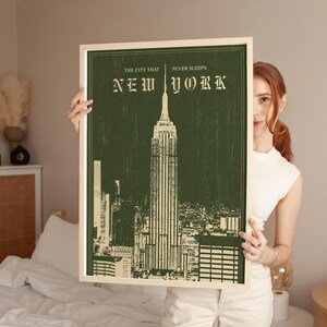 May include: A rectangular framed wall art print featuring a dark green background with a cream-colored illustration of the Empire State Building. The text at the top reads THE CITY THAT NEVER SLEEPS and NEW YORK in a decorative serif font.