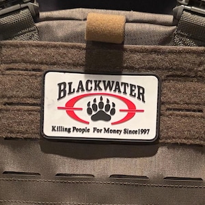 May include: A rectangular morale patch attached to a brown tactical vest. The patch features a white background with a black bear paw print inside a red oval. The text reads BLACKWATER at the top and Killing People For Money Since 1997 below.