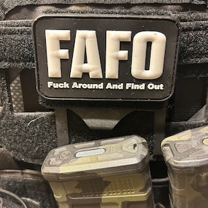 May include: A black tactical vest with a black patch that reads "FAFO" in white letters and "Fuck Around And Find Out" in smaller white letters. Two camouflage-patterned magazine pouches are attached to the vest.